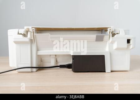 White multifunction printer with exposed power cable and battery on a wooden desktop, clean modern office workspace ready for printing, scanning and e Stock Photo