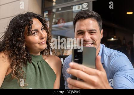 Happy young couple enjoying drinks together and taking a selfie with a smartphone at an outdoor restaurant terrace. Casual lifestyle moment with beer, Stock Photo