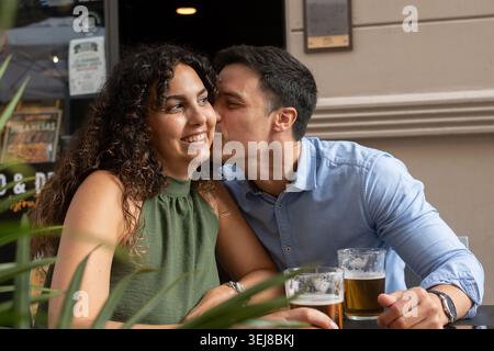 Happy young couple enjoying a romantic moment together at an outdoor restaurant terrace. Man kissing woman on the cheek while sharing beers during a r Stock Photo