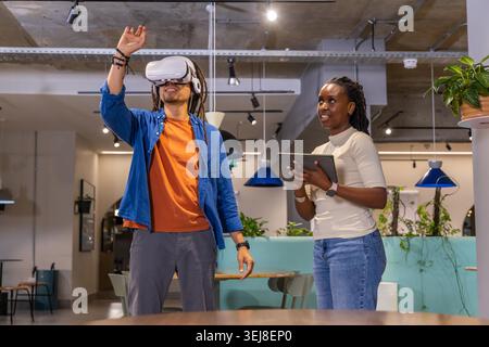 Diverse coworkers demoing VR, man wearing orange tee reaching up, woman holding tablet in office. Coworking, collaboration, headset, gesture, virtual, interface, modern Stock Photo