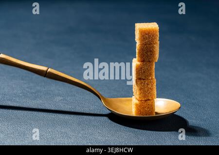 Tower of raw brown sugar cubes in a golden spoon over a blue textured surface, close up. Minimalist concept for sweetness portion control and diet Stock Photo