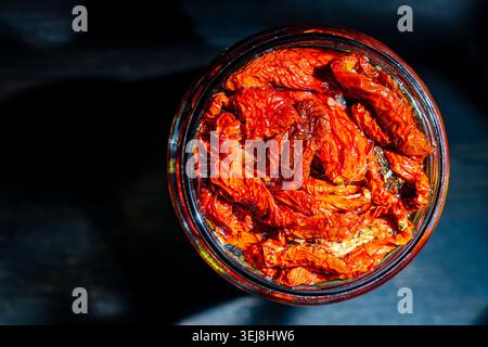 Gourmet sun dried tomatoes in oil inside a glass jar on dark background, close up. Top view of traditional Mediterranean food appetizer with moody lig Stock Photo