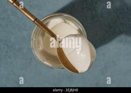 Top view of thick white yogurt or cream cheese or sour cream in a glass jar with a golden spoon on a blue background. Minimalist healthy breakfast con Stock Photo