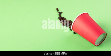 Coffee beans spilling from a red disposable cup on a vibrant green background, perfect for concepts of caffeine, waste, or refreshment. panoramic imag Stock Photo