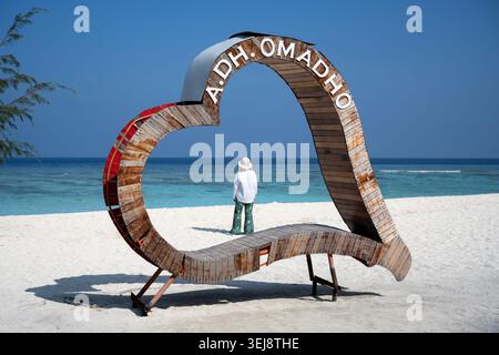 A photograph of a tropical beach featuring a large, rustic wooden heart-shaped structure resting on white sand. The structure has the white text "A.DH Stock Photo