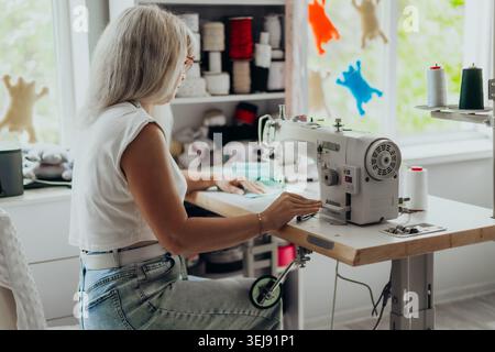 Woman with gray hair sewing fabric on a professional sewing machine in a bright modern studio. Creative workspace with colorful threads and handmade Stock Photo