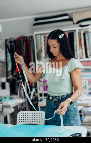 Confident female fashion designer measuring fabric in a modern sewing studio, surrounded by textiles and creative tools. She is focused on precision Stock Photo