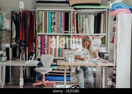 Fashion designer working in a creative studio filled with colorful fabrics and sewing materials. Woman sewing at a professional machine, surrounded by Stock Photo