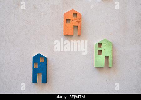 Three colorful, wooden house cutouts in blue, orange, and green are displayed on a textured gray wall. Each cutout features square windows and a doorw Stock Photo