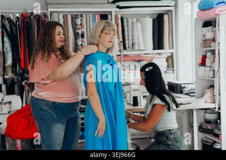 Fashion designers collaborate in a creative studio, fitting a vibrant blue dress on a mature woman model. Tailoring, teamwork, and modern style trends Stock Photo