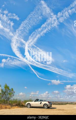 Silver convertible sports car parked on a rural hilltop beneath dramatic aircraft contrails and cirrus clouds in a blue sky. Stock Photo