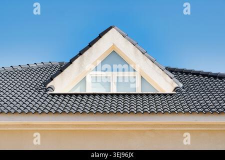 Triangular dormer window on a black tiled roof of a modern house against a clear blue sky. Stock Photo