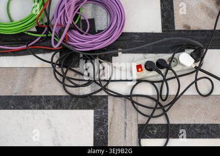 A white power strip with plugged-in black cords is lying at an indoor marble floor setting. Stock Photo