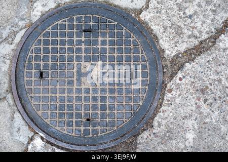 Circular cast iron manhole cover with the letter K is embedded in cracked concrete pavement. Stock Photo