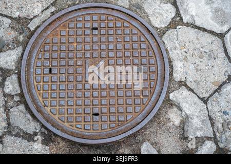 Rusted cast iron manhole cover marked with the letter K sits flush within cracked concrete and stone pavement. Stock Photo