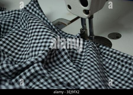 Industrial sewing machine stitching black and white checkered fabric in a garment manufacturing unit. Close-up view of textile production. Stock Photo