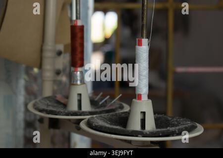 Close-up of textile machine spindles holding white and red yarn cones in a weaving workshop, highlighting thread feeding mechanisms. Stock Photo