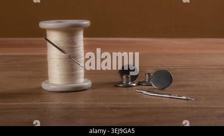 Spool of white sewing thread with needles, snap fasteners and sewing tools arranged on a wooden surface, representing tailoring, garment making and textile craftsmanship. Stock Photo