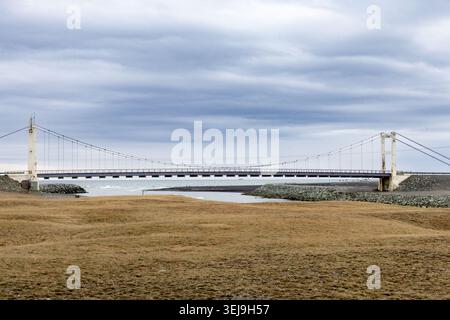 Suspension bridge crossing Icelandic coastal river under dramatic cloudy Nordic sky Stock Photo