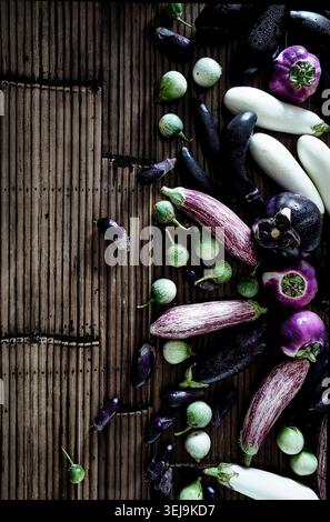 Top view of assorted eggplants arranged on a rustic bamboo mat background. The composition features a variety of aubergines in deep purple, white, and Stock Photo