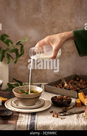 pumpkin soup in a bowl on a wooden table, with roasted hazelnuts and sage leaves, someone is pouring fresh cream into the soup Stock Photo