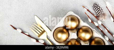 Elegant Easter flat lay with golden eggs in carton, willow branches, feathers, bunny figurine and gold cutlery on light stone background. Festive spri Stock Photo