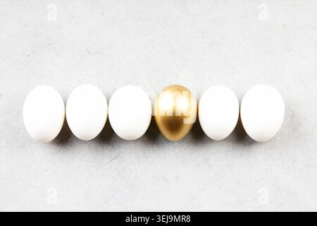 Modern minimal Easter composition with one golden egg standing out among white eggs on light stone background. Clean spring concept with copy space. Stock Photo