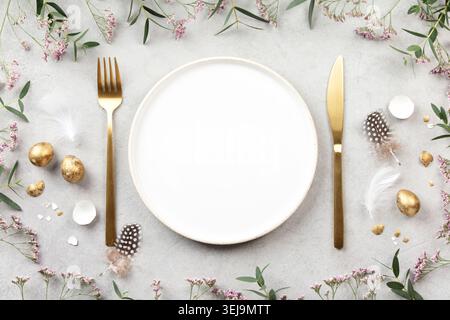 Elegant Easter table composition with empty white plate, golden cutlery, gold eggs, feathers and pink spring flowers on light stone background. Festiv Stock Photo