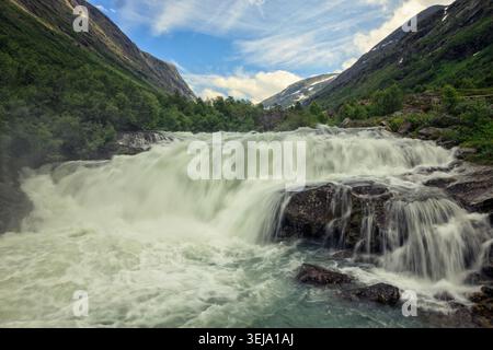 Gamle Strynefor Norway - 28.06.2024: A long-exposure shot of a rushing waterfall flowing over dark rocks in a verdant mountain valley, showcasing the Stock Photo