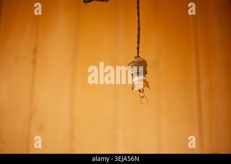 An old, damaged brass light bulb socket with a shattered glass bulb base hangs vertically from a dusty cord. Stock Photo