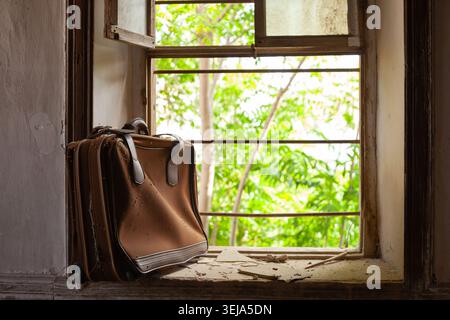 A single aged brown suitcase with leather trim handles leans upright against the sill of a deteriorated, iron-barred window in an abandoned room. Stock Photo