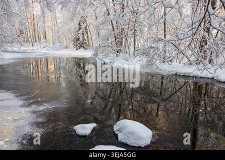 Gumböle, Finland - 31.12.2023: Partially frozen river in a snowy winter forestA dark, flowing river with patches of ice winds through a dense forest, Stock Photo