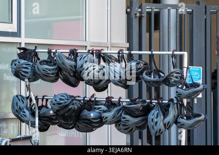 Rows of bicycle helmets hang from a metal rack outside a city building. The image shows cycling safety gear, rental equipment, and urban mobility. Stock Photo