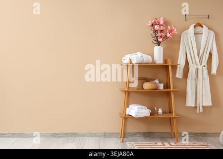 Shelf unit with artificial flowers, towels and soft bathrobe on beige wall in bathroom Stock Photo