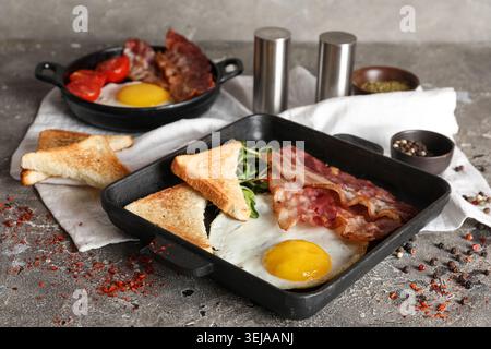 Two portions of fried eggs, bacon, toasts and bowls of spices on table against grey background Stock Photo