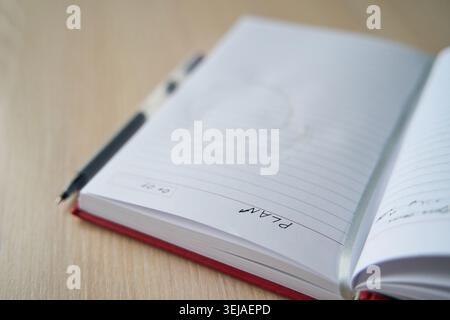 Red Notebook Opened Showing Lined Page With Handwritten Plan Note And Pen Placed On Wooden Table In Shallow Depth Of Field Workspace Scene With Soft Blur. Stock Photo