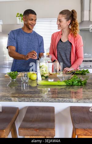 Diverse couple in blue shirt, jacket using blender and chopping greens on granite island in kitchen Stock Photo