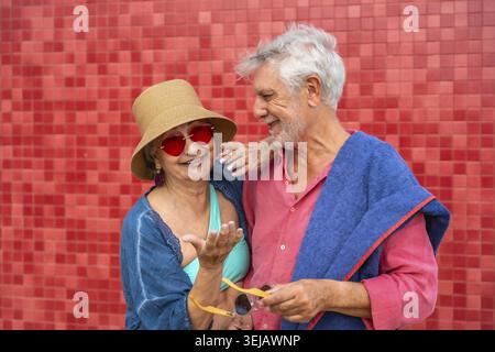 Happy senior couple standing against a red tiled wall, sharing a relaxed moment with smiles and wearing summer attire, symbolizing an active and joyfu Stock Photo