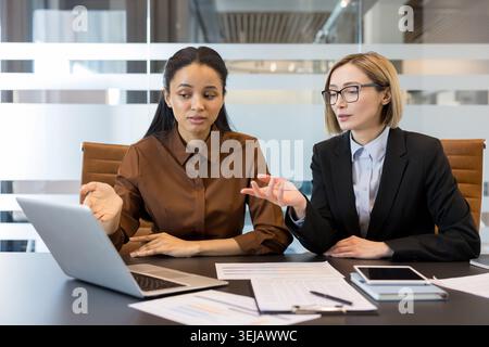 Diverse businesswomen discussing a project while collaborating on a laptop in a modern office, showing teamwork, communication, and professional partnership during a meeting Stock Photo