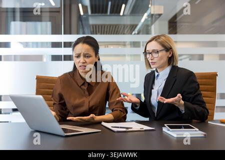 Two stressed businesswomen sitting at a conference table, looking at a laptop screen with expressions of confusion and frustration, discussing a problem or error during an office meeting Stock Photo