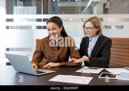 Two diverse businesswomen smiling and collaborating on a laptop, discussing work and sharing ideas in a contemporary office setting, fostering teamwork and mentorship Stock Photo