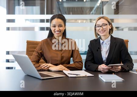 Two diverse businesswomen smiling confidently at the camera while sitting at a modern office desk with a laptop, tablet, and documents, representing success and teamwork in a corporate environment Stock Photo