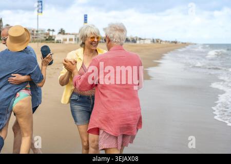Senior couples laughing and dancing together on a sunny sandy beach, enjoying an active retirement vacation by the ocean, sharing joy, connection, and Stock Photo