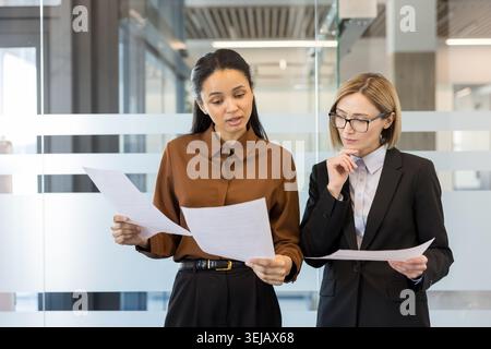 Two diverse businesswomen reviewing paperwork and communicating in a modern office, fostering teamwork and professional collaboration while analyzing business strategies Stock Photo