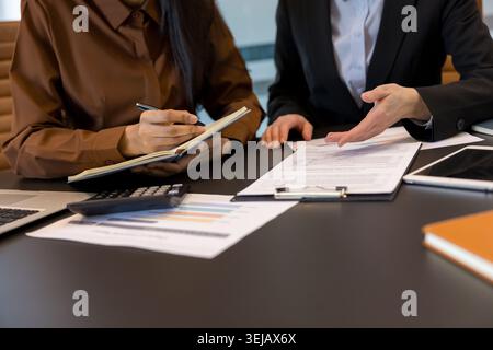 Two businesswomen review financial reports and collaborate over paperwork, taking notes and analyzing charts at a desk with laptop, calculator and notebooks in a modern office Stock Photo