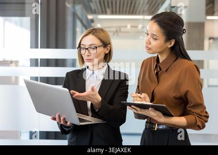 Two multiracial professional business women standing in an modern office actively discussing a new business project, while one holds an open laptop and the other takes notes Stock Photo