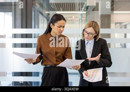 Two diverse businesswomen standing in an office hallway, holding papers and looking stressed, discussing complex financial data on documents with expressions of frustration and disagreement Stock Photo