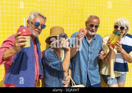 Group of smiling senior friends holding refreshing drinks and glazed donuts, celebrating an active summer vacation together against a bright yellow ba Stock Photo