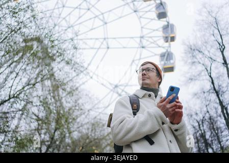 Young man with backpack and smartphone is standing near ferris wheel in amusement park, looking away and using mobile phone, wearing warm white jacket Stock Photo