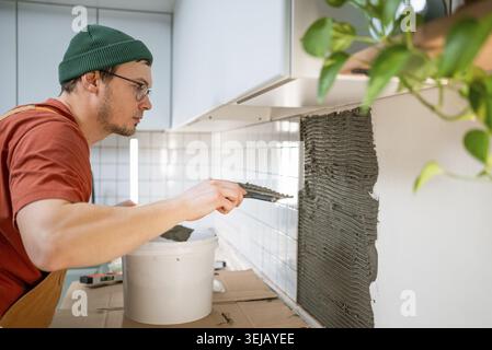 Tiler applying tile adhesive onto a kitchen wall, preparing for ceramic backsplash installation and home renovation, focusing on the details of home i Stock Photo